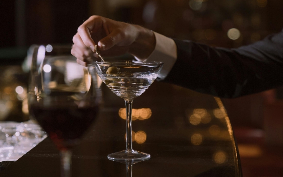 a man holding a martini olive at a cocktail bar with a glass of red wine sitting next to it